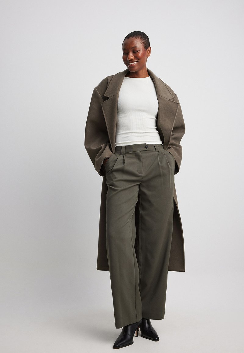 Olijfgroene wijde broek met hoge taille, gecombineerd met een getailleerde witte tanktop en een lange bruine jas, afgewerkt met zwarte haklaarzen.