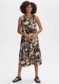 Floral print sleeveless dress in beige and black, tiered design, featuring a round neckline and relaxed fit. Black sandals complete the look.