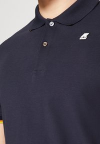 Navy polo triko s texturovanou látkou, dvěma knoflíky a stříbrnou logem na hrudi. Límec má kontrastní akcent.