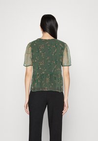 Grön blommig peplum-blus med korta transparenta ärmar, prydd med ett delikat mönster och en krökt nederkant, kombinerad med svarta vida byxor.