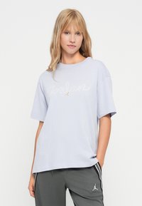 Jonge vrouw met blond haar, gekleed in een lichtpaars Jordan logo t-shirt en donkergrijze sportbroek met witte strepen en logo.