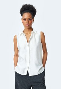 Weiße ärmellose Bluse mit Kragen und durchgehender Knopfleiste, aus strukturiertem Stoff. Kombiniert mit gestreiften dunklen Hosen.