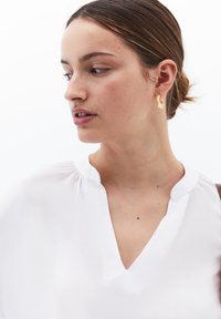 Blusa bianca leggera con scollatura a V e dettagli arricciati sulle spalle; modella che indossa orecchini dorati asimmetrici.