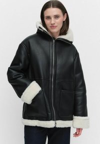 Svart konstläderjacka med en vit pälsfoderhuva och manschetter, med två framfickor och dragkedja.