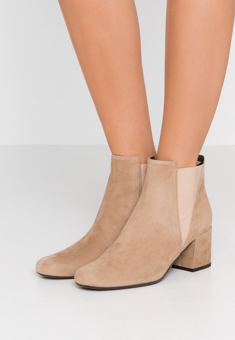Femme portant des bottines beiges en daim avec talons carrés, panneaux latéraux élastiques, debout sur un sol blanc devant un fond uni.