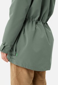 Giacca verde leggera con vita arricciata, texture liscia e tasche laterali. Include un piccolo logo verde sull'orlo.