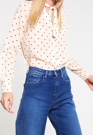 Kvinde iført cremefarvet bluse med lange ærmer med brune prikker og blå højtsiddende jeans, stående mod en ensfarvet hvid baggrund.