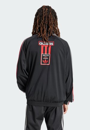 Zwarte sportjack met rode accenten, voorzien van een logo-opdruk en verticale strepen op de achterkant. Zacht, textuurstof, relaxte pasvorm.