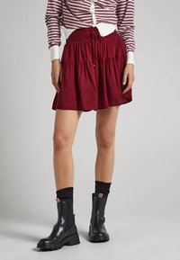 Kvinna i röd lagerkjol, randig långärmad tröja, svarta ankelskor och svarta strumpor som står mot en grå bakgrund.