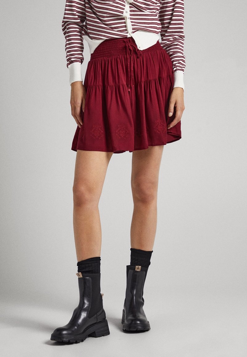 Kvinna i röd lagerkjol, randig långärmad tröja, svarta ankelskor och svarta strumpor som står mot en grå bakgrund.