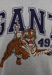 Sweat-shirt gris avec un grand tigre brodé en orange et noir, aux yeux verts, au-dessus d'un grand texte en gras disant "GANT 49."