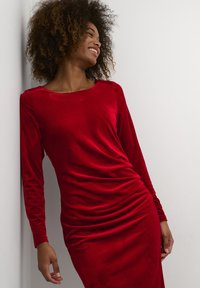 Robe en velours rouge à manches longues avec un décolleté bateau et des détails froncés sur le côté, présentant une texture douce et une silhouette ajustée.