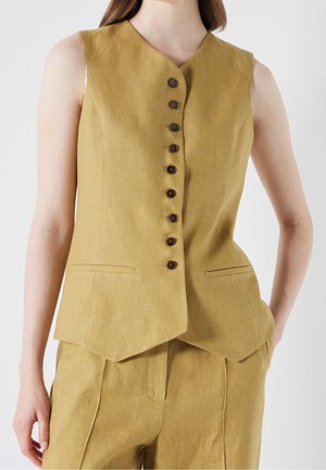 Vrouw draagt een mosterdgele mouwloze button-down vest en bijpassende broek, staand met één hand in haar zak tegen een effen achtergrond.