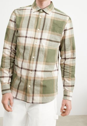 Mann trägt ein langärmeliges, kariertes Hemd in Grün, Beige und Weiß mit Brusttasche und weiße Hose vor einfarbigem Hintergrund.