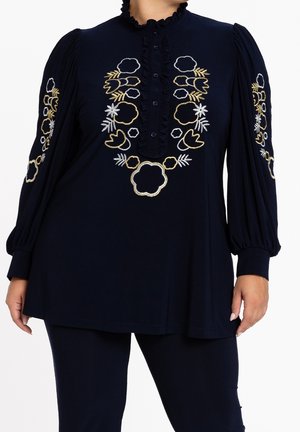 Mannequin grande taille portant une tunique bleu marine avec col volanté, patte de boutonnage et broderie florale or et blanche sur le devant et les manches.