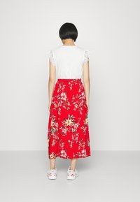 Top blanco con detalles de encaje en los hombros, combinado con una falda larga roja con flores, que presenta flores amarillas y rosas, usada con zapatillas blancas.