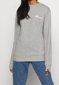 Femme portant un sweat-shirt gris ellesse et un jean bleu, debout devant un fond clair uni.