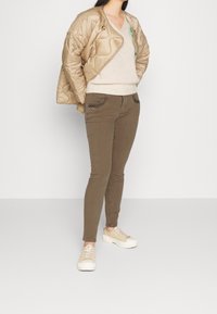 Chaqueta acolchada color beige claro, suéter crema de cuello en V, jeans ajustados marrones con bolsillos texturizados y zapatillas beige con suela gruesa.