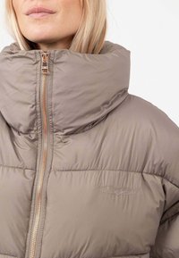 Giacca a vento in beige chiaro con colletto alto, chiusura con zip e logo sottile a forma di ala. Presenta una morbida texture trapuntata, ideale per il calore.