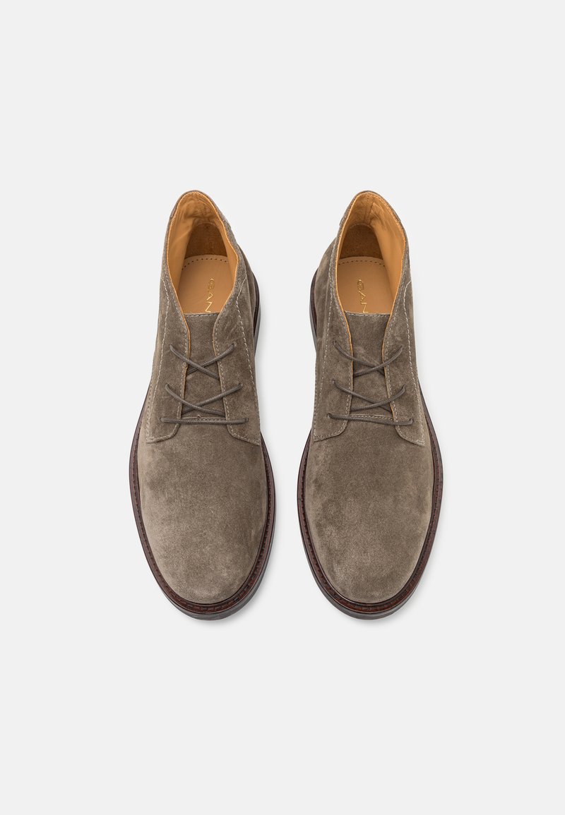 Para brązowych zamszowych butów na sznurówki z skórzaną podszewką i ciemnobrązowymi podeszwami, widziana z góry na białym tle.