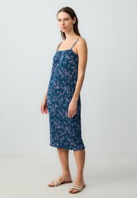 Ärmelloses Midikleid in dunkeltürkis mit floralem Muster. Verfügt über ein plissiertes Design und einen gewellten Saum. Getragen mit beigen Riemchensandalen.