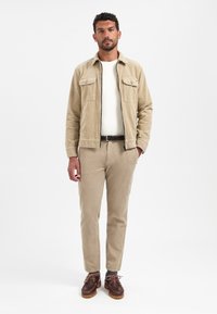 Beige Corduroy-Jacke mit zwei Brusttaschen, kombiniert mit passenden gerippten Hosen und braunen Schnürschuhen, vor einem weißen Hintergrund.