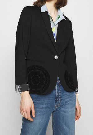Sort tætsiddende blazer med enkelt knap, cirkulær blomsterbroderi på lommerne, båret over en skjorte med blomsterprint og blå jeans.