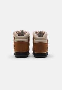 Bottines en cuir marron avec un col rembourré beige, semelles en caoutchouc noir et un logo sur le côté extérieur. Finition texturée et design robuste.