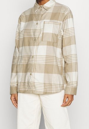 Person trägt ein beige-weiß kariertes Hemd mit Knöpfen, Brusttasche und langen Ärmeln, kombiniert mit weißen Hosen.