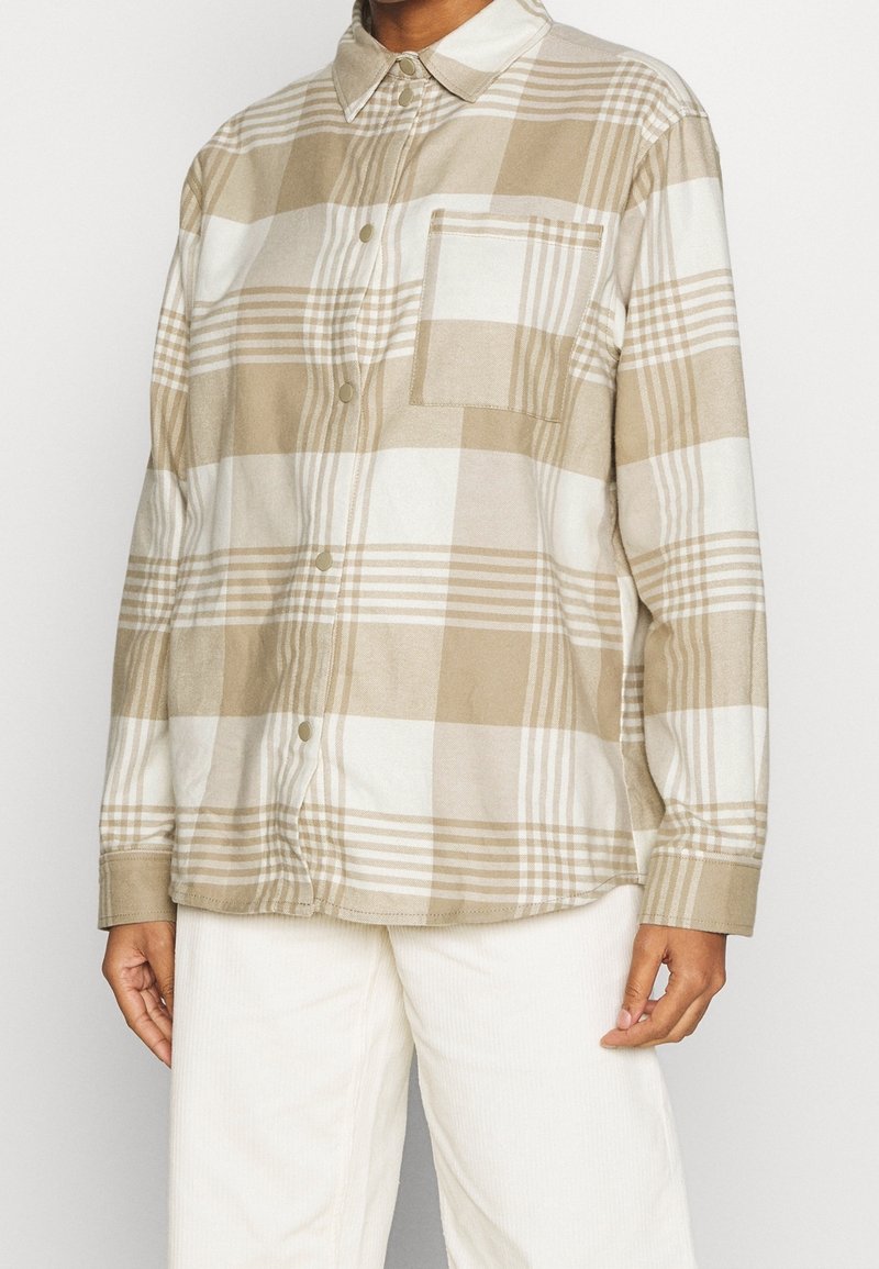 Personne portant une chemise à grands carreaux beige et blanc, à boutons, avec une poche poitrine et des manches longues, associée à un pantalon blanc.