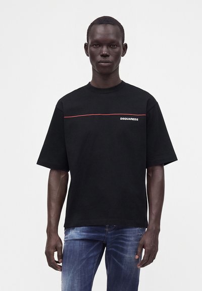 Homme portant un t-shirt noir Dsquared2 avec un détail de ligne rouge, associé à un jean bleu, debout devant un fond clair uni.