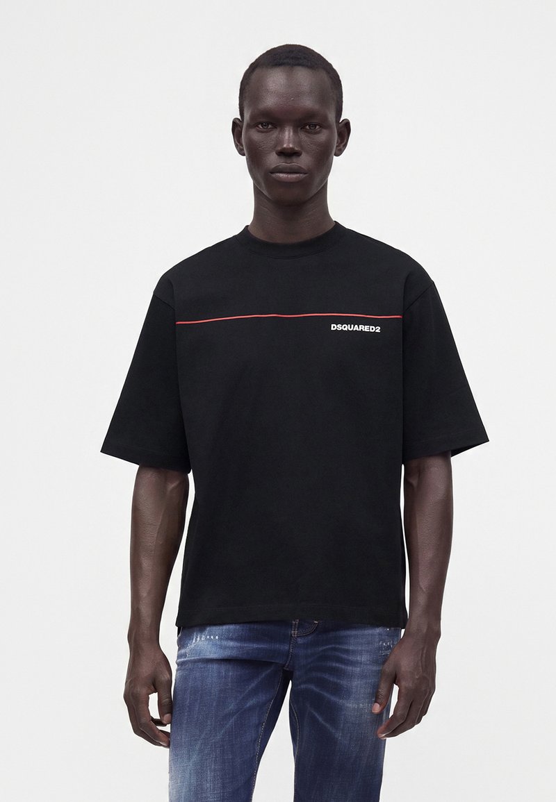 Άνδρας που φοράει μαύρο t-shirt Dsquared2 με κόκκινη λεπτομέρεια γραμμής, συνδυασμένο με μπλε τζιν, στέκεται μπροστά σε απλό φωτεινό φόντο.