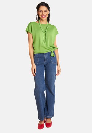 Lächelnde Frau, die ein grünes kurzärmliges Oberteil mit Seitenknoten, eine grüne Perlenkette, blaue Jeans und rote Schuhe trägt und vor einem weißen Hintergrund steht.