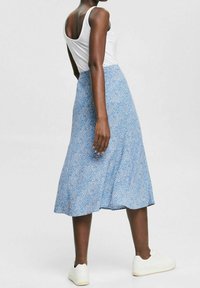 Mujer vestida con una blusa sin mangas blanca, una falda midi azul con estampado y zapatillas blancas, de pie contra un fondo claro y liso.