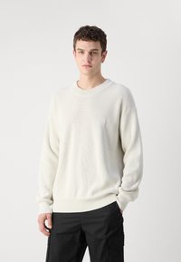 Maglione bianco a coste con scollatura rotonda e logo discreto sul petto, dotato di maniche lunghe e vestibilità comoda, abbinato a pantaloni neri.