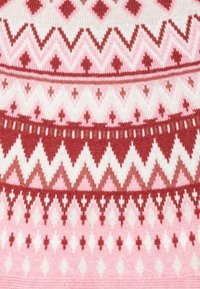 Stickad tyg med ett geometriskt mönster som har växlande rader av röda, rosa och vita diamanter och zickzackar. Mjuk textur och varma färger.