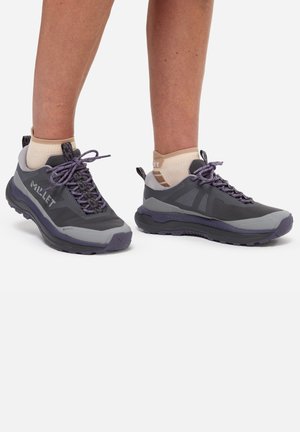 Personne portant des chaussures de randonnée Millet grises et violettes et des socquettes beige, debout sur un fond blanc.