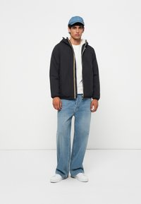 Veste rembourrée noire avec capuche et fermeture éclair dorée, portée sur une chemise blanche ; assortie à un jean large bleu clair et des baskets blanches.