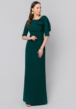 Kobieta ubrana w długa suknię w ciemnozielonym kolorze z rękawami do łokci i dużą kokardą na lewym ramieniu, stojąca na szarym tle.