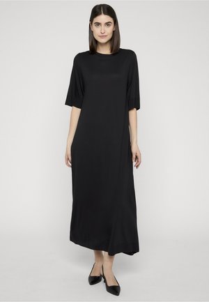 Kobieta o prostych ciemnych włosach nosi luźną czarną sukienkę midi z rękawami do łokcia oraz czarne buty na obcasie ze zapięciem na piętę i szpiczastym noskiem.