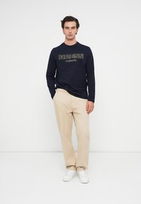 Camiseta de manga longa em azul-marinho com o logótipo "GUESS" impresso, combinada com calças chino bege e ténis brancos, contra um fundo branco liso.