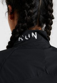 Henkilö, jolla on tummat palmikkohiuset, pukeutuneena mustaan takkiin, jossa on osittain näkyvää heijastavaa tekstiä kauluksessa, valkoista taustaa vasten.