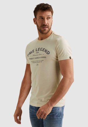 Man met kort bruin haar die een beige T-shirt met print van PME Legend en een blauwe spijkerbroek draagt, licht naar rechts kijkend.