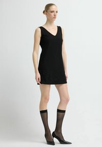 Czarna sukienka bez rękawów z dekoltem w serek, ozdobiona koralikami na dole. Modelka ma na sobie czarne, sięgające kolan skarpetki i szpilki z ostrym noskiem.