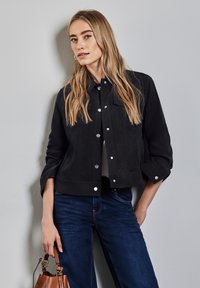 Svart mockajacka med kort design, prydd med silverknappar och uppvikta ärmar, kombinerad med mörkblå jeans och en brun handväska.