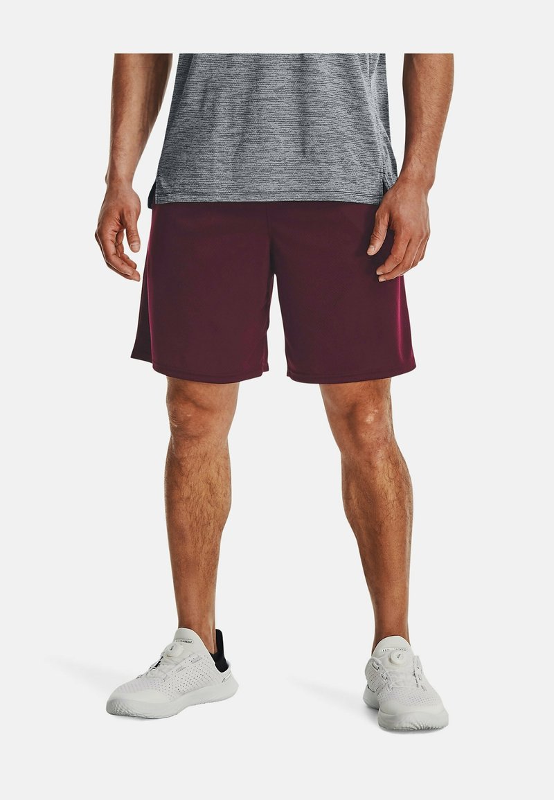 Homme portant un short de sport bordeaux, un t-shirt gris et des baskets blanches, debout devant un fond uni.