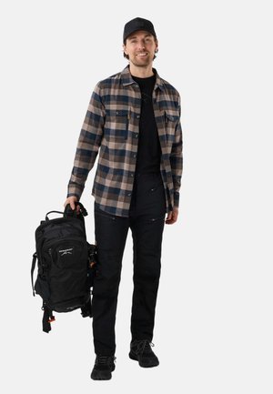 Smiling man in black cap, plaid shirt, black pants, and shoes holds black Swedmount backpack by side, standing on plain white background.