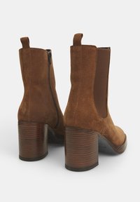 Bottines en daim marron avec talon bloc. Présentent des panneaux élastiques latéraux et une fermeture éclair arrière pour faciliter l'enfilage. Talon en bois texturé.