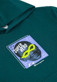 Felpa teal con un patch grafico colorato che presenta del testo, un volto sorridente e occhiali gialli. Il materiale sembra morbido e lavorato a maglia.
