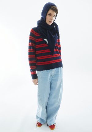 Persoon in een marineblauw en rood gestreept trui, lichtblauwe wijde jeans, marineblauwe sjaal om het hoofd gewikkeld, en rode sneakers, staand tegen een witte achtergrond.